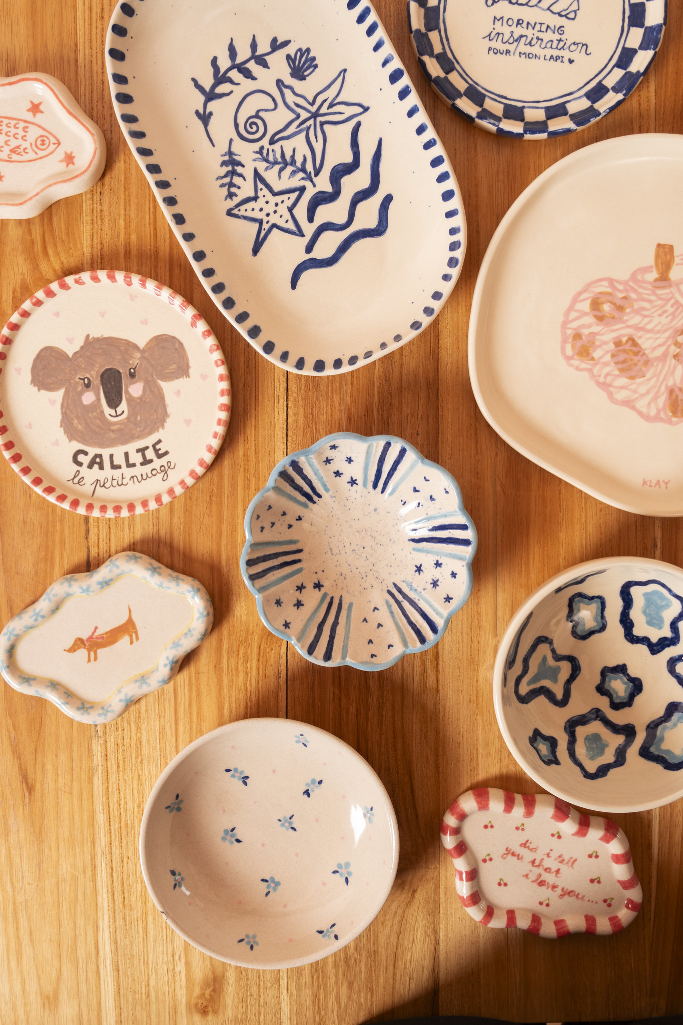 Hand-painted ceramic plates, dishes, and mugs laid out on a wooden table at Klay Bar in Canggu, showcasing the varied patterns and personal designs created during painting sessions.